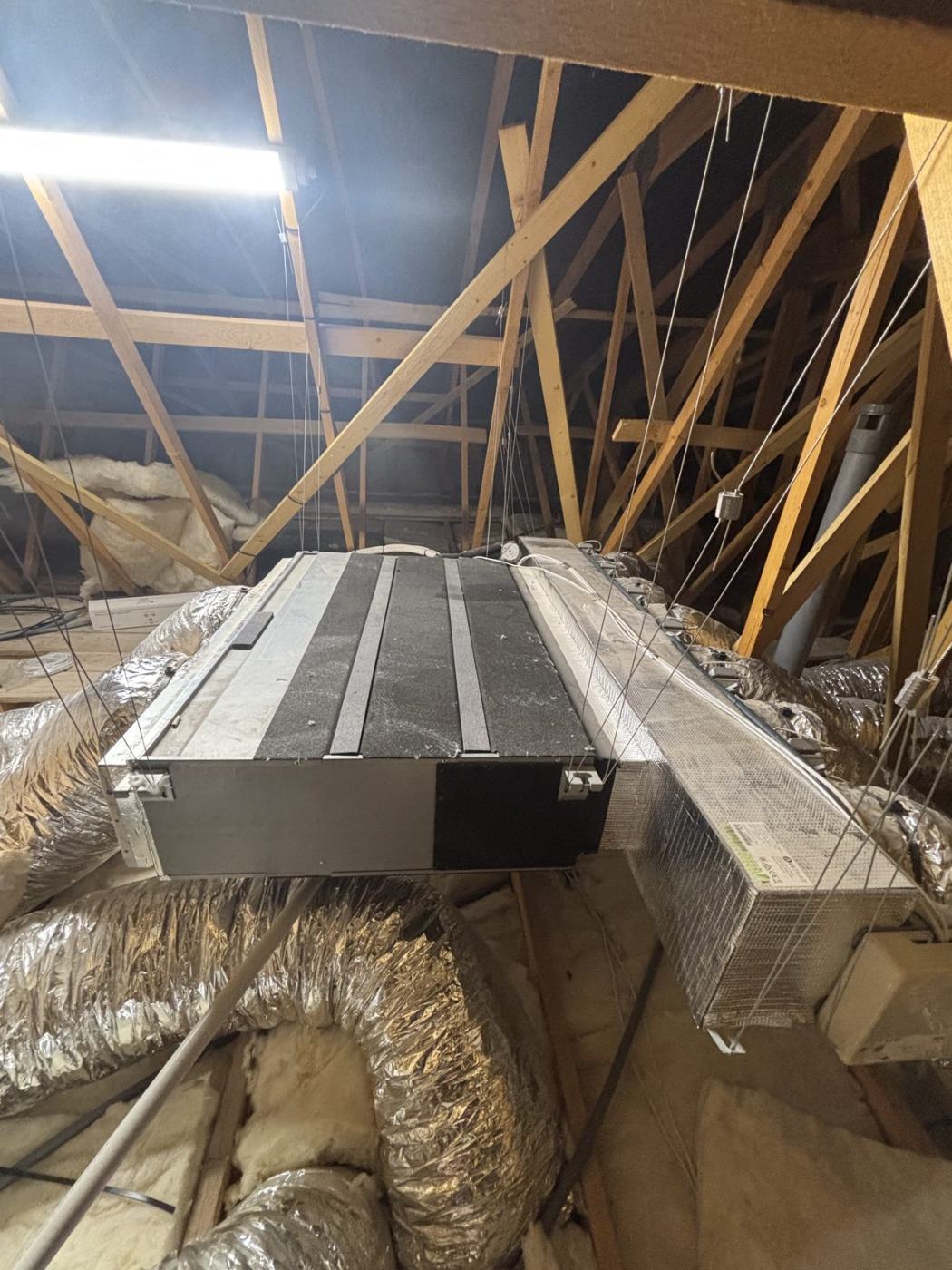 Unité de climatisation gainable suspendue par câbles sous une charpente en bois, avec son plénum et ses gaines isolées